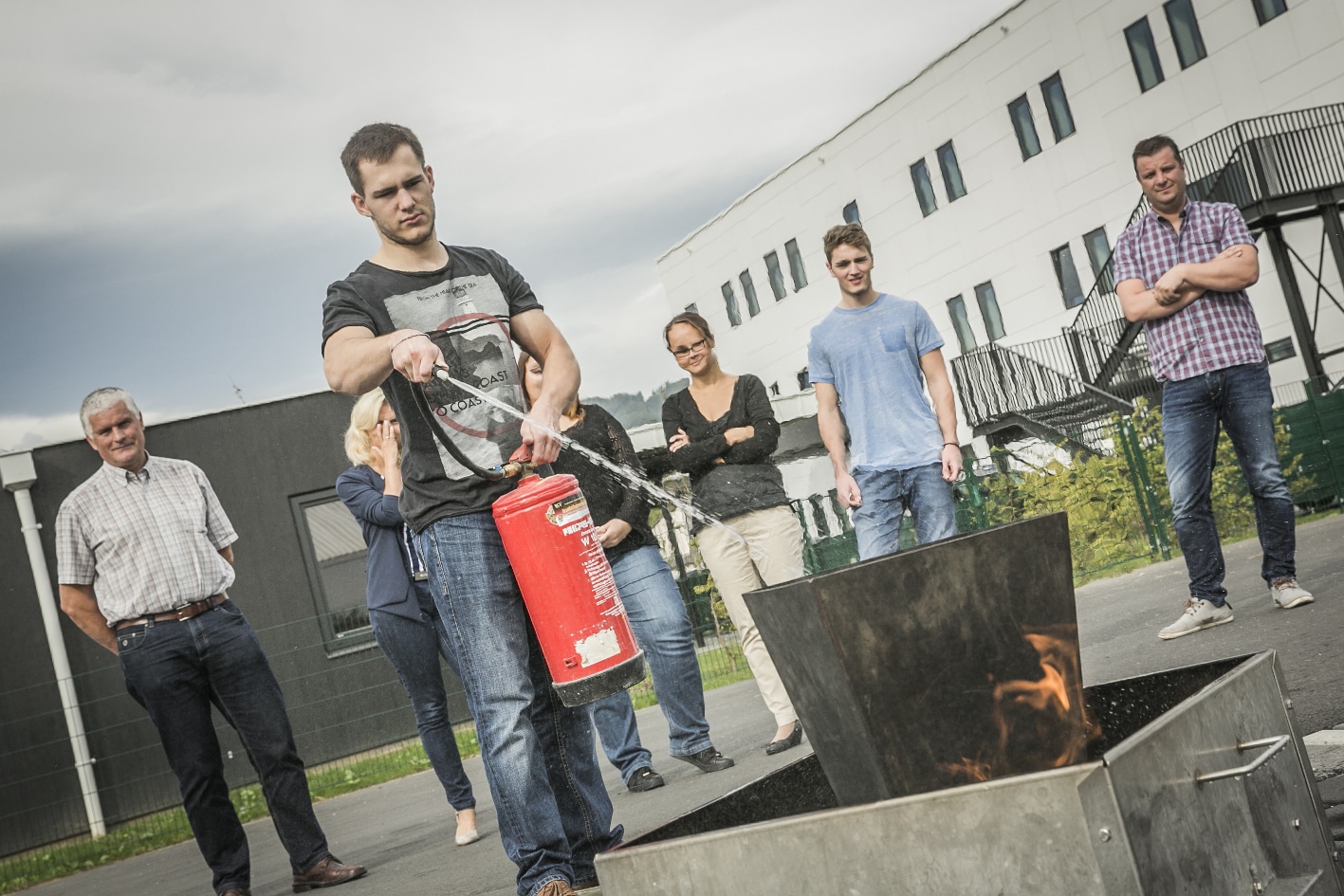 Das Bild zeigt einen Mann, der mit einem Handfeuerlöscher ein kleines Feuer in einem Eimer löscht. Hinter ihm stehen Menschen und schauen zu.